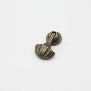 May include: A bronze-toned metal drawer pull featuring a circular mounting base and a hinged, fan-shaped handle with vertical ridges. The hardware has a weathered, dark finish with highlights on the raised edges of the fan-shaped pull component.