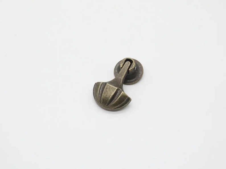 May include: A bronze-toned metal drawer pull featuring a circular mounting base and a hinged, fan-shaped handle with vertical ridges. The hardware has a weathered, dark finish with highlights on the raised edges of the fan-shaped pull component.