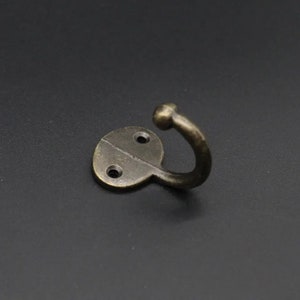 Könnte beinhalten: Ein bronzefarbener Metallhaken mit abgerundeter Oberseite und flachem Boden. Der Haken hat zwei Schraubenlöcher an der Basis.