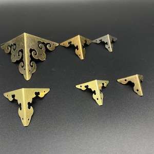 May include: Assortment of six gold metal corner brackets with ornate designs. The brackets are of varying sizes and are arranged on a black surface.