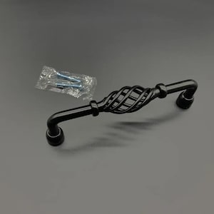 May include: A matte black cabinet pull featuring a central twisted cage design with two smooth arms extending to circular mounting bases. The hardware includes two silver-colored metal screws enclosed in a small, clear plastic bag for installation.