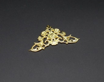 May include: A gold-colored metal corner bracket featuring an intricate, symmetrical filigree design with scrolling patterns and small circular holes for mounting. The piece has a pointed top and two extended arms, showcasing a polished finish.