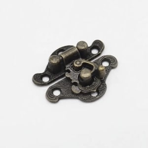 May include: Antique bronze-colored clasp with ornate floral detailing. The clasp features a hinged design with a decorative latch and two mounting plates with screw holes. The metal has a weathered appearance.