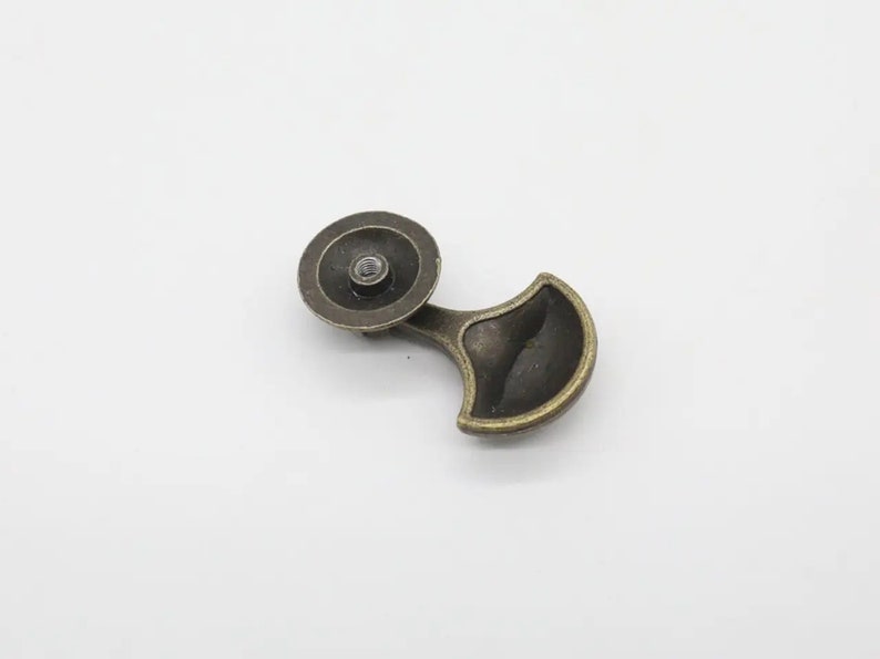 May include: A bronze-toned metal drawer pull featuring a circular mounting base with a central threaded hole and a curved, fan-shaped handle extension. The hardware has a weathered, dark metallic finish with visible highlights on the edges.