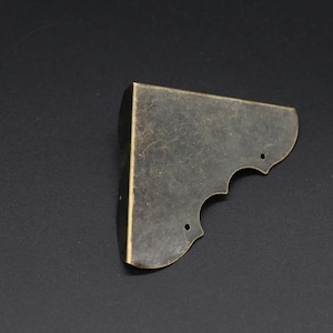 Puede incluir: Un solo soporte de esquina de latón antiguo con un borde decorativo festoneado. El metal tiene un acabado envejecido y dos pequeños agujeros para el montaje. El soporte está diseñado para reforzar la esquina de una caja o marco.