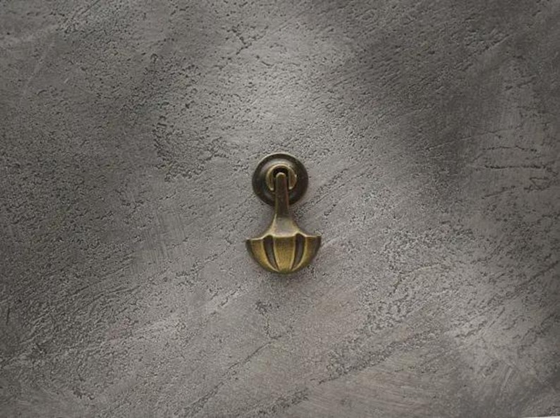 May include: A brass-toned metal drop pull handle featuring a circular backplate and a fan-shaped pendant. The hardware has a weathered, antique finish with visible vertical ridges on the lower section of the drop handle component.