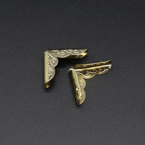 May include: Two L-shaped metal corner protectors with a polished gold-tone finish. Each piece features an embossed scrollwork pattern along the outer edges and a smooth, curved design intended for securing the corners of books or boxes.