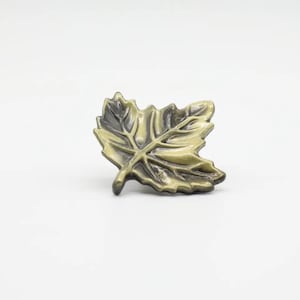 Może przedstawiać: Metalowy gałka do szafki w kształcie liścia, w kolorze złotym i czarnym.