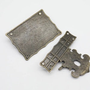 May include: A two-piece antique brass-toned metal latch set. The rectangular plate features scalloped edges and four mounting holes. The corresponding latch component has a decorative, ornate shape with a central opening and a small metal pin.