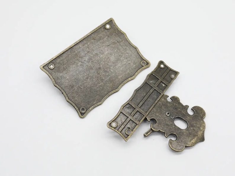 May include: A two-piece antique brass-toned metal latch set. The rectangular plate features scalloped edges and four mounting holes. The corresponding latch component has a decorative, ornate shape with a central opening and a small metal pin.