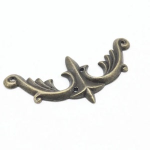 May include: A decorative metal accent piece in an antique brass finish. The piece is shaped like a stylized double-headed bird with intricate details and two holes.