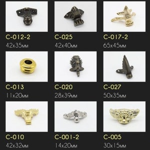 Op de afbeelding: Assortiment van decoratieve metalen hoekbeugels in verschillende stijlen en maten. De beugels zijn gemaakt van goud, zilver en zwart metaal en hebben ingewikkelde ontwerpen. De beugels zijn gelabeld met hun bijbehorende codes en afmetingen in millimeters.