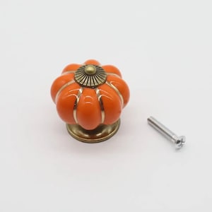 May include: Orange ceramic pumpkin-shaped cabinet knob with a gold-toned metal base and a screw for installation.