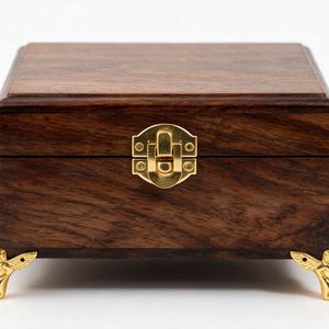 May include: A rectangular dark brown wooden box with a visible wood grain pattern. The box features a polished gold-colored metal latch on the front center and four decorative gold-colored metal feet shaped like small winged figures.