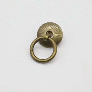 May include: A bronze ring pull cabinet handle with a decorative, circular, fluted top.