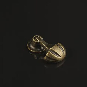 May include: A brass-toned metal furniture pull with an antique finish. The hardware features a circular base plate connected to a hinged, semi-circular handle with three vertical grooves, designed for attachment to a drawer or cabinet door.