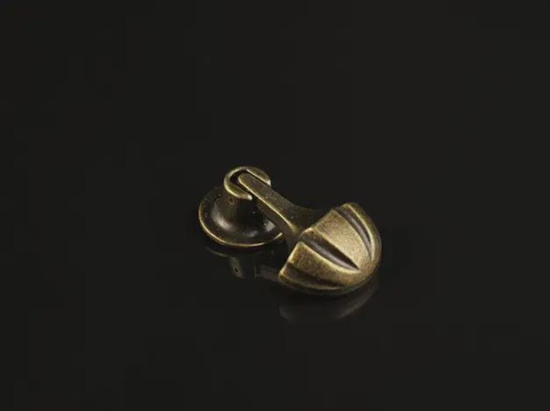 May include: A brass-toned metal furniture pull with an antique finish. The hardware features a circular base plate connected to a hinged, semi-circular handle with three vertical grooves, designed for attachment to a drawer or cabinet door.