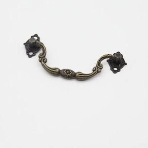 Może przedstawiać: Dekoracyjny metalowy uchwyt do ciągnięcia w kolorze brązu z ozdobnymi detalami. Uchwyt ma zakrzywiony kształt i dwie kwadratowe płytki z otworami na śruby.