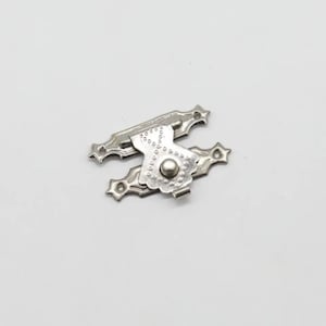 Könnte beinhalten: Ein silberfarbener Metall-Kistenverschluss mit einer dekorativen, verzierten Grundplatte mit sternförmigen Enden und einem zentralen Schwenkarm. Der Arm hat ein texturiertes Punktmuster und einen kleinen runden Knopf zum Greifen und Sichern.