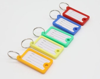 May include: A collection of colorful key tags in red, blue, green, yellow, and orange. Each tag has a silver key ring and a white label with lines for name, address, city, and country. The tags are arranged diagonally.