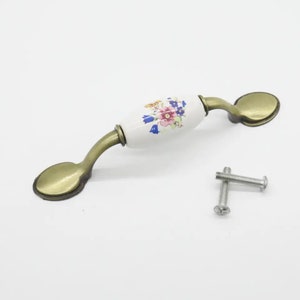 May include: A cabinet pull with a white ceramic center adorned with a floral pattern in pink, blue, and purple. The handle has a curved design with bronze-colored metal accents and two screws.