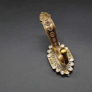 Op de afbeelding: Een gouden decoratieve haak met een bloemmotief en heldere strass steentjes. De haak is ovaalvormig met een puntig uiteinde.