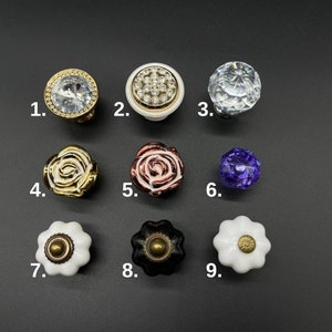May include: Nine different cabinet knobs in various styles and colours. The knobs are made of ceramic, glass, and metal. They feature designs such as roses, diamonds, and geometric patterns.