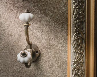 May include: A wall-mounted metal hook featuring an antique brass finish and two white ceramic knobs. The hook has a curved arm design with a circular mounting plate and is positioned next to a decorative frame with intricate scrollwork detailing.