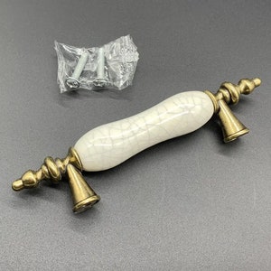 Puede incluir: Un tirador de cajón con un cuerpo central de cerámica color blanco roto con acabado craquelado oscuro, flanqueado por dos soportes de metal en tono latón antiguo con bases cónicas y extremos en forma de remate, con dos tornillos.