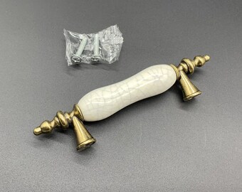 Può includere: Una maniglia per cassetto con un corpo centrale in ceramica bianco sporco con finitura craquelé scura, affiancata da due supporti in metallo color ottone antico con basi coniche e terminali a punta, inclusa una coppia di viti.