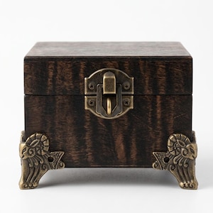 May include: A dark brown wooden box with a visible wood grain pattern. The box features a central brass-colored metal latch and four decorative brass-colored metal feet shaped like ram heads at each corner of the base of the square container.