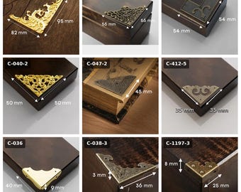 May include: A collection of nine metal corner protectors for boxes and books, featuring various gold, bronze, and brass finishes with intricate patterns. Each piece is labeled with a code and includes visible measurements in millimeters.