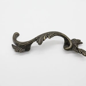 Può includere: Pomello per armadio ornato color bronzo con un design curvo. La maniglia presenta dettagli di foglie e volute, aggiungendo un'estetica vintage. Ideale per il restauro di mobili o per aggiungere un tocco decorativo.