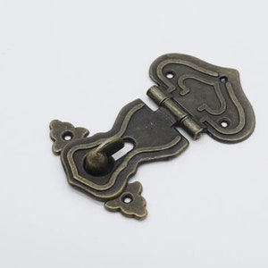 May include: Antique brass latch with a decorative design and a hook. The latch has a hinge and is designed for use on a wooden box or door.