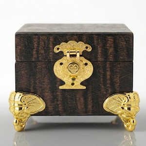 May include: A dark brown wooden box with a visible wood grain pattern. The box features a decorative gold-colored metal latch on the front and four gold-colored metal feet shaped like elephant heads with prominent ears and trunks.
