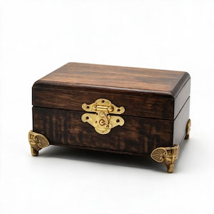 May include: A rectangular dark brown wooden box featuring a polished gold-tone metal latch on the front. The box is supported by four ornate gold-tone metal feet, each shaped like an elephant head with detailed ears and a trunk facing outward.