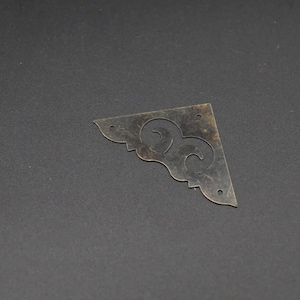 May include: A triangular metal corner protector with a dark, weathered brass finish. The piece features a decorative cutout pattern of two swirling curves and includes three small circular holes for mounting purposes along its straight edges.