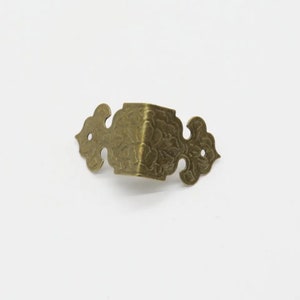 May include: A brass-toned metal corner protector featuring an ornate, embossed floral pattern. This hardware component has two symmetrical, curved wings extending from a central angled corner, each containing a small circular hole for mounting.