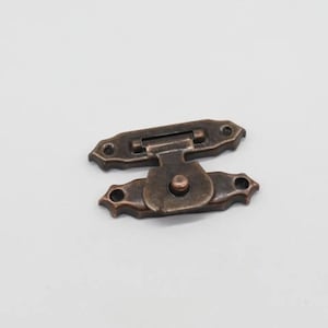 Puede incluir: Un cierre de metal de estilo vintage, de color cobre antiguo, con un diseño decorativo. El cierre tiene un pequeño pomo redondo y un mecanismo de bisagra.