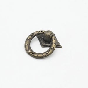 Könnte beinhalten: Ein bronzener Ring-Schrank-Knopf mit einem dekorativen Design. Der Knopf ist an einer quadratischen Basis befestigt.