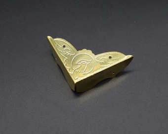 May include: A gold-colored metal corner protector featuring an engraved bird motif within a circular frame and decorative scrollwork. The L-shaped hardware includes two small pre-drilled holes on each side for attachment to a box or furniture edge.