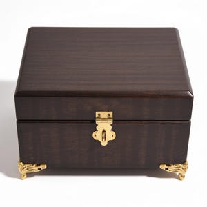 May include: A square dark brown wooden box with a smooth finish. The box features a polished gold-coloured metal latch on the front centre and four ornate gold-coloured metal feet at the base corners, providing a stable and elevated appearance.