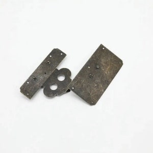 Op de afbeelding: Drie antiek ogende metalen onderdelen voor een kast scharnier. De onderdelen zijn rechthoekig en hebben een verweerde afwerking. De onderdelen zijn met schroeven verbonden.
