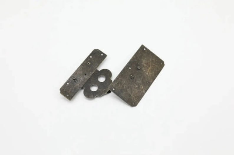 Op de afbeelding: Drie antiek ogende metalen onderdelen voor een kast scharnier. De onderdelen zijn rechthoekig en hebben een verweerde afwerking. De onderdelen zijn met schroeven verbonden.