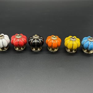 以下が含まれることがあります： 白、赤、黒、オレンジ、黄、青など、さまざまな色のセラミック製かぼちゃ型の引き出しノブが6個あります。各ノブには金色の金属ベースが付いています。