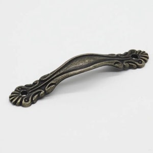May include: An antique-style cabinet pull handle with a curved design. The handle is made of metal with an aged bronze finish. Decorative floral scrollwork adorns the ends of the handle, which has two mounting holes.