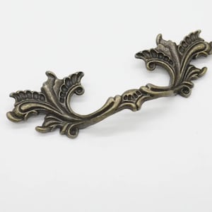 Op de afbeelding: Een sierlijke, antieke, bronzen kleurige kastknop met een decoratief, bloemig ontwerp.