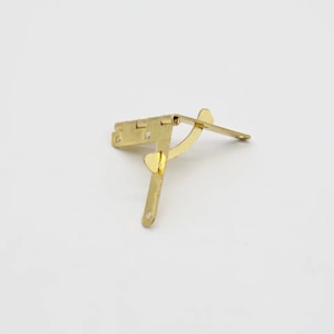 May include: A gold-toned metal hinge with a curved arm and a small, round knob. The hinge is designed for attaching two surfaces at a 90-degree angle.
