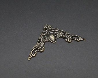 May include: A triangular metal corner protector with an antique bronze finish, featuring intricate scrollwork and leaf-like patterns. The piece includes a central teardrop-shaped indentation and small circular holes at the ends for attachment.