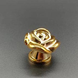 May include: A gold-coloured rose-shaped cabinet knob with white accents. The knob has a detailed floral design and a round base. The hardware is likely for furniture or decorative purposes.